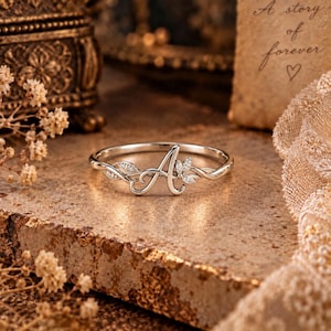 May include: A silver-toned ring featuring a prominent letter A as the centrepiece, flanked by delicate leaf-shaped accents embedded with small, clear stones. The band is thin and polished, showcasing a refined design with intricate metalwork.