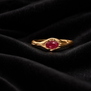 Könnte beinhalten: Ein Goldring mit einem zentralen, oval geschliffenen roten Edelstein in einer Krappenfassung. Die Ringschiene weist eine texturierte, organische Oberfläche auf, die zur Mitte hin schmaler wird und den leuchtend roten Stein sicher im Gold einfasst.