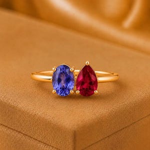 Può includere: Un anello in oro con due pietre preziose prominenti affiancate. Una pietra è una gemma blu a taglio ovale, l'altra è una gemma rossa a taglio a goccia. L'anello è in metallo dorato lucido e le pietre sono fissate da piccole griffe in oro.