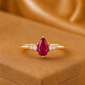 Può includere: Un anello in oro con una pietra preziosa rossa centrale a forma di pera. La fascia è accentuata su ogni lato da tre piccole pietre bianche rotonde e trasparenti incastonate nell'oro. Il design è simmetrico, con la pietra al centro.