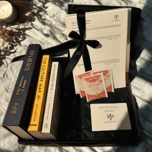 Peut inclure: Un coffret cadeau noir contenant des livres, un agenda et des sachets de thé. Les livres incluent "Influence", "The Culture Map" et "Surrounded by Idiots". L'agenda porte l'inscription "Week View". L'ensemble est noué avec un ruban noir.