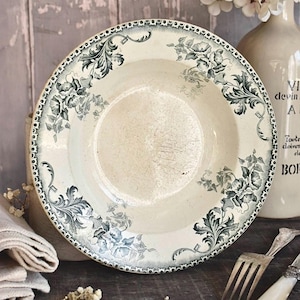 Puede incluir: Un plato redondo de cerámica con base color crema y un patrón de transferencia verde oscuro con motivos florales y hojas de acanto. El borde está decorado con un diseño festoneado repetitivo y el centro muestra un ligero desgaste superficial.