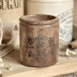 May include: A cylindrical brown ceramic crock with a crackled glaze finish. The front features a black printed label that reads J. SAINSBURY'S, SUPERIOR, HOME MADE, and POTTED MEATS, centered around a large letter S inside a circular emblem.