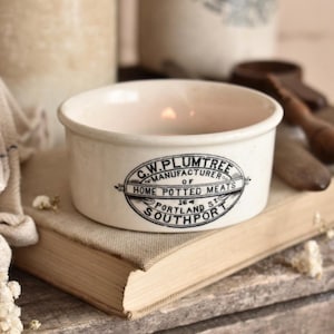 May include: A round, off-white ceramic bowl featuring a black printed label that reads G.W. PLUMTREE MANUFACTURER OF HOME POTTED MEATS 164 PORTLAND ST SOUTHPORT. The bowl has a smooth, cylindrical rim and a shallow interior basin.