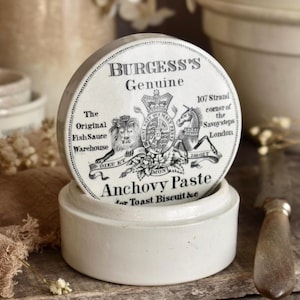 Può includere: Un vasetto rotondo in ceramica bianca con coperchio caratterizzato da testo stampato in nero e uno stemma reale. Il testo recita Burgess's Genuine Anchovy Paste, The Original Fish Sauce Warehouse, 107 Strand corner of the Savoy steps London, e for Toast Biscuit &e.