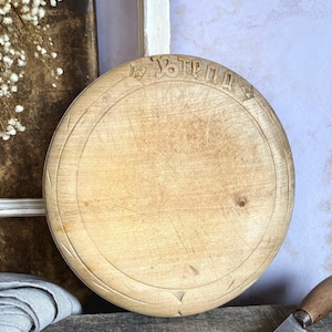 Puede incluir: Una tabla de pan redonda de madera clara con un borde tallado y la palabra "BREAD" inscrita en una fuente decorativa en la parte superior. La superficie muestra un desgaste visible, incluyendo numerosos rasguños y marcas de cuchillo.