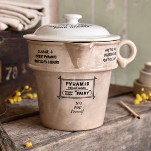 Può includere: Uno scaldavivande in ceramica beige con coperchio, caratterizzato da scritte nere: 'Clarke's Patent Pyramid Night Lights 9 Hours', 'Pyramid Trade Mark Fairy', 'No 3 Pint' e 'Price 6/-'. Il contenitore presenta un manico laterale e un beccuccio.