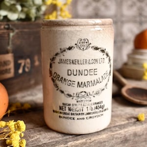 May include: A cylindrical ceramic jar with a cream-coloured crackle glaze finish. The front features black printed text reading James Keiller & Son Ltd. Dundee Orange Marmalade, with a net weight of 1 lb. (454g) and origin details.