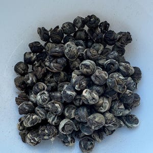 May include: A close-up of a collection of tea balls, each tightly wound and dark green with white accents. The tea balls are clustered together in a white bowl, showcasing their intricate, floral-like appearance.