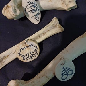 Könnte beinhalten: Nahaufnahme mehrerer Knochenanhänger mit dunkelblauen Sigill-Designs. Die Anhänger sind mit Metallschlaufen an Knochenfragmenten befestigt. Der Hintergrund ist eine dunkelblaue Oberfläche.