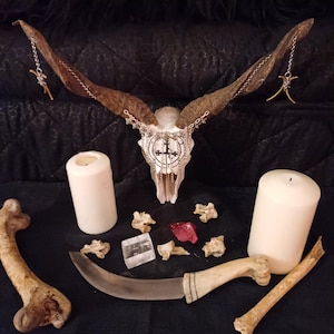 May include: A decorative arrangement featuring a goat skull with large horns, adorned with a cross pendant and chain. The scene includes two white candles, a curved knife, bones, and crystals, all set against a dark background.
