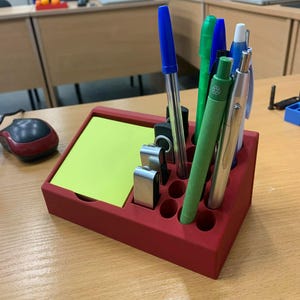 May include: A red desk tidy with a section for sticky notes and several holes for pens and pencils. The tidy holds various pens and pencils in blue, green, and silver. A yellow sticky note pad is in the tidy.