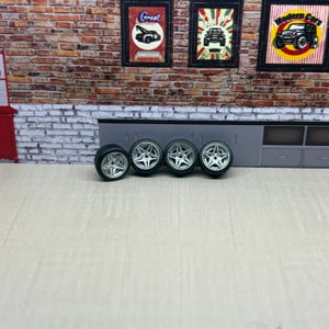May include: Four silver-colored car wheels with a five-spoke design. The tires are black. The wheels are displayed on a light-colored surface in front of a brick wall with framed car-themed artwork.