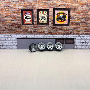 Puede incluir: Cuatro ruedas de modelo de coche negras y plateadas con letras blancas, dispuestas en fila. Las ruedas se colocan frente a una pared de ladrillo adornada con carteles de automóviles enmarcados. La escena evoca un garaje o taller.