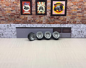 May include: Four black and silver model car wheels with white lettering, arranged in a row. The wheels are set against a backdrop of a brick wall with framed automotive-themed posters. The scene evokes a garage or workshop setting.