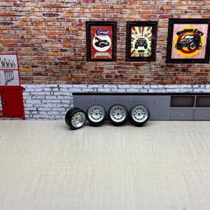 May include: Four black and silver model car wheels are displayed against a backdrop of a brick wall and framed automotive-themed posters. The wheels have a silver rim and black tires, suggesting a focus on automotive detailing or model car customization.