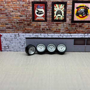May include: Four black and silver toy car wheels with a detailed silver rim design. The wheels are set against a backdrop of a brick wall and framed vintage car posters, suggesting a car enthusiast theme.