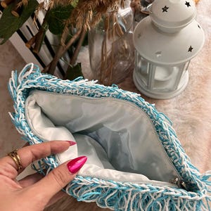 May include: A handmade clutch with a blue and white fringe design. The bag has a zipper closure and a light blue interior. The clutch is made of a woven material and is open, revealing the inside. A white lantern with star cutouts is in the background.