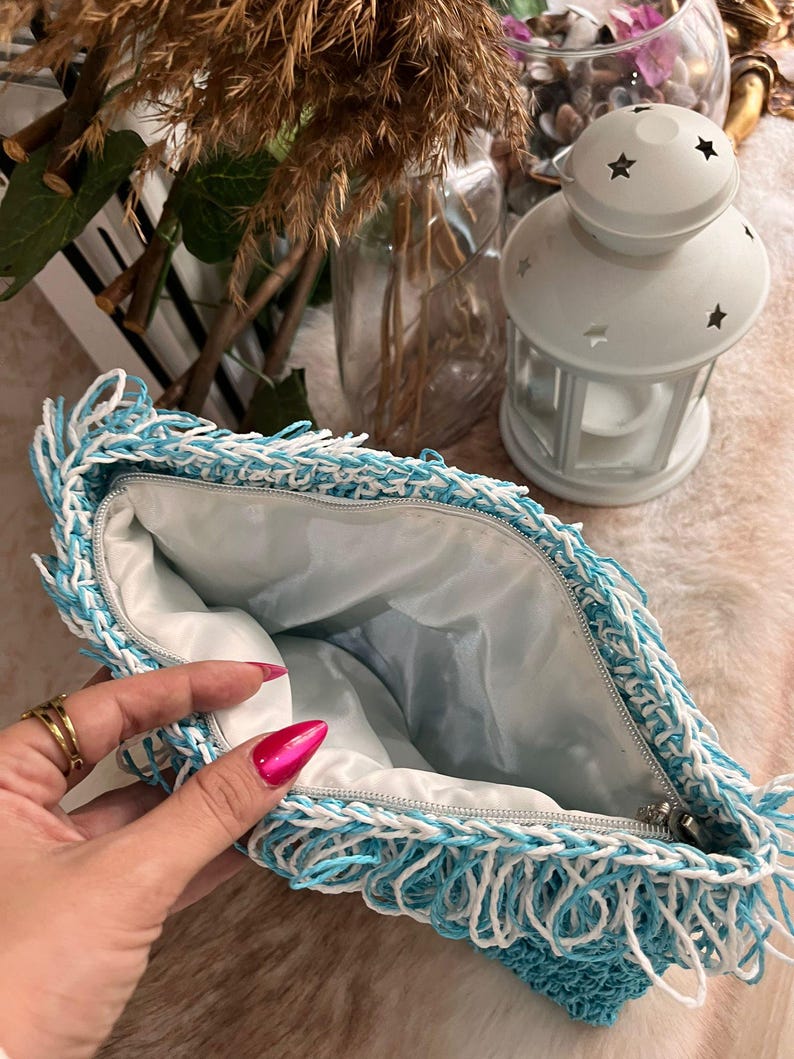 May include: A handmade clutch with a blue and white fringe design. The bag has a zipper closure and a light blue interior. The clutch is made of a woven material and is open, revealing the inside. A white lantern with star cutouts is in the background.