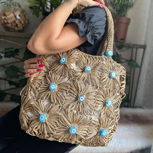 May include: A beige tote bag with a floral design made of woven material. The flowers have light blue centers. The bag has a woven handle and is held by a person.