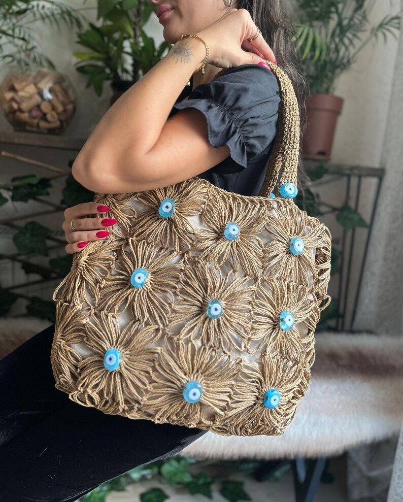 May include: A beige tote bag with a floral design made of woven material. The flowers have light blue centers. The bag has a woven handle and is held by a person.