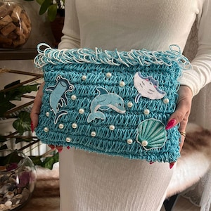 May include: A turquoise crocheted clutch with white fringe and pearl accents. The clutch features embroidered sea creatures, including a dolphin, a stingray, a frog, and a seashell. The bag is held by a person.
