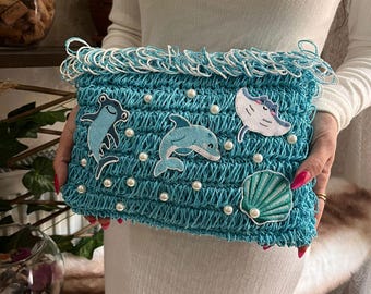 Könnte beinhalten: Eine türkisfarbene gehäkelte Clutch mit weißem Fransen und Perlenakzenten. Die Clutch zeigt gestickte Meeresbewohner, darunter ein Delfin, ein Rochen, ein Frosch und eine Muschel. Die Tasche wird von einer Person gehalten.