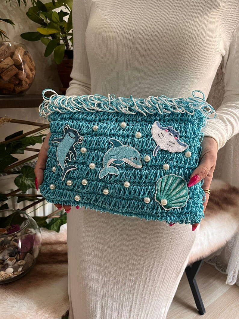 May include: A turquoise crocheted clutch with white fringe and pearl accents. The clutch features embroidered sea creatures, including a dolphin, a stingray, a frog, and a seashell. The bag is held by a person.