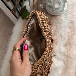 K&ouml;nnte beinhalten: Eine gewebte Stroh-Clutch mit goldenem Rei&szlig;verschluss und schillernden Pailletten. Der Innenraum ist mit einem Satin-Stoff gef&uuml;ttert. Die Clutch ist offen und gibt den Blick ins Innere frei. Die Tasche ist hellbraun.