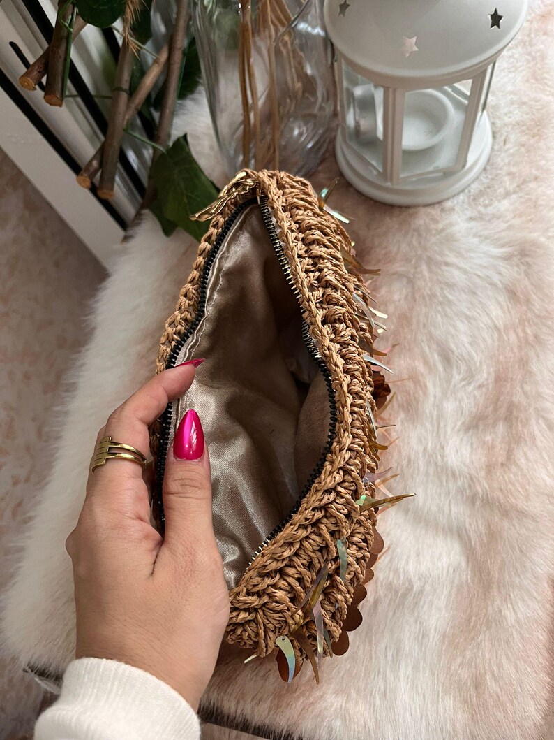 K&ouml;nnte beinhalten: Eine gewebte Stroh-Clutch mit goldenem Rei&szlig;verschluss und schillernden Pailletten. Der Innenraum ist mit einem Satin-Stoff gef&uuml;ttert. Die Clutch ist offen und gibt den Blick ins Innere frei. Die Tasche ist hellbraun.