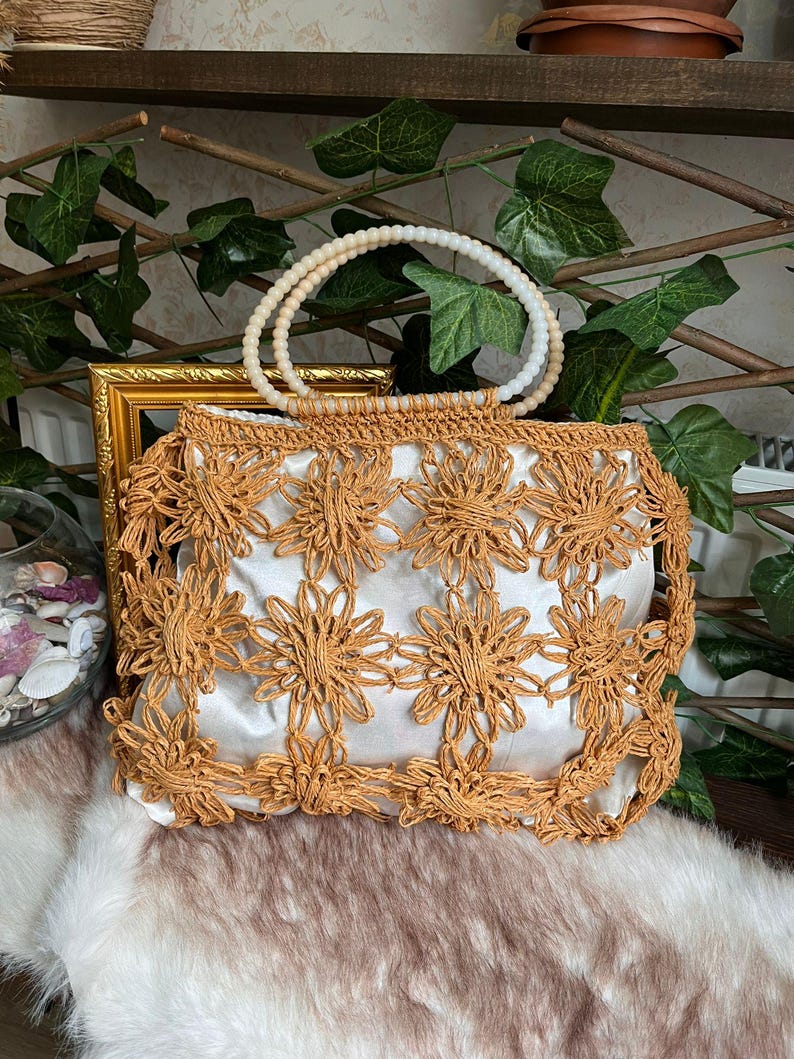 May include: A tan-colored, open-weave handbag with a white satin lining. The bag features a floral design and a beaded handle. The bag is sitting on a faux fur surface.