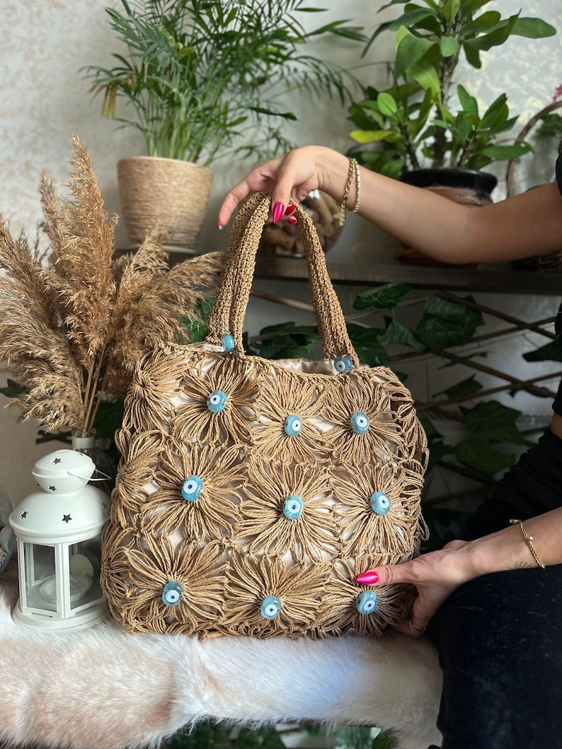 May include: A woven tote bag with a floral design. The bag is made of tan-colored woven material and features a pattern of tan flowers with blue centers. The handles are also tan and woven. The bag is held by a person.