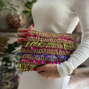 May include: A woven raffia clutch with decorative accents. The rectangular bag features layers of brown raffia, embellished with green, purple, and iridescent fringe, and pink sequin accents. The clutch is held by a person wearing a white dress.