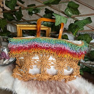 Puede incluir: Un bolso tote hecho a mano con un asa de madera. El bolso presenta una franja de arco&iacute;ris en la parte superior y un dise&ntilde;o floral en la parte delantera. El bolso est&aacute; hecho de materiales naturales y tiene un interior blanco.