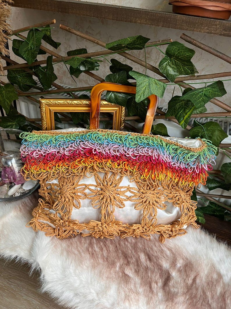 Puede incluir: Un bolso tote hecho a mano con un asa de madera. El bolso presenta una franja de arco&iacute;ris en la parte superior y un dise&ntilde;o floral en la parte delantera. El bolso est&aacute; hecho de materiales naturales y tiene un interior blanco.