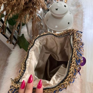 May include: Open view of a gold-colored chain bag with blue and gold fringe. The bag has a gold-colored chain strap and a beige satin interior. The zipper is black. The bag is on a white fur-like surface.