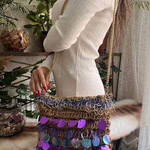 May include: A woven shoulder bag with a gold chain strap. The bag features a brown base, blue fringe, and rows of large, iridescent purple sequins. The bag is a stylish accessory.