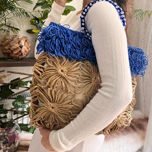 May include: A handmade tote bag with a unique design. The bag features a beige, flower-like woven pattern on the front and a blue fringe detail. The strap is adorned with blue and white beads. The bag is held by a person wearing a white long-sleeved shirt.