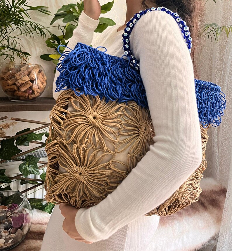 May include: A handmade tote bag with a unique design. The bag features a beige, flower-like woven pattern on the front and a blue fringe detail. The strap is adorned with blue and white beads. The bag is held by a person wearing a white long-sleeved shirt.
