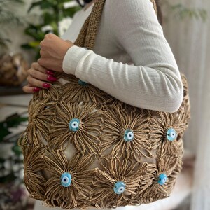 May include: A beige tote bag with a floral design made of woven material. The flowers have light blue eye-shaped embellishments in the center. The bag has a woven handle and is held by a person.