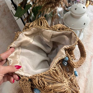 May include: A woven straw tote bag with a beige satin lining. The bag features a fringe design and is adorned with blue eye-shaped beads. The handles are also made of woven straw. The bag is open, revealing the interior.