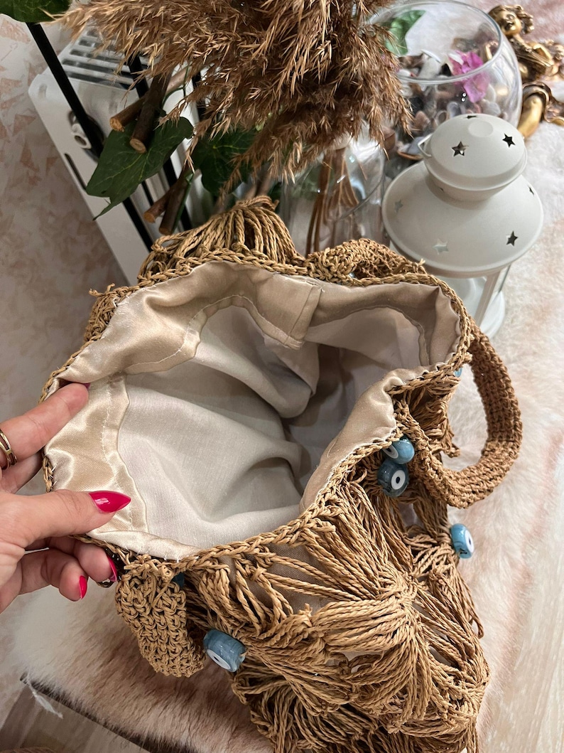 May include: A woven straw tote bag with a beige satin lining. The bag features a fringe design and is adorned with blue eye-shaped beads. The handles are also made of woven straw. The bag is open, revealing the interior.