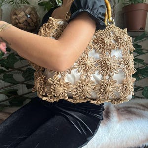K&ouml;nnte beinhalten: Eine beige Handtasche mit floralem H&auml;keldesign. Die Tasche hat einen Bambusgriff und ein wei&szlig;es Innenfutter. Die Tasche wird von einer Person getragen, die ein schwarzes Oberteil und eine schwarze Hose tr&auml;gt.