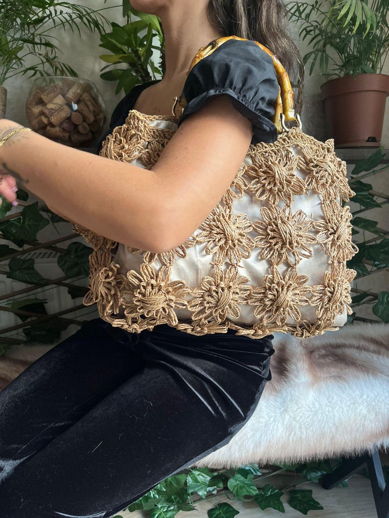 K&ouml;nnte beinhalten: Eine beige Handtasche mit floralem H&auml;keldesign. Die Tasche hat einen Bambusgriff und ein wei&szlig;es Innenfutter. Die Tasche wird von einer Person getragen, die ein schwarzes Oberteil und eine schwarze Hose tr&auml;gt.