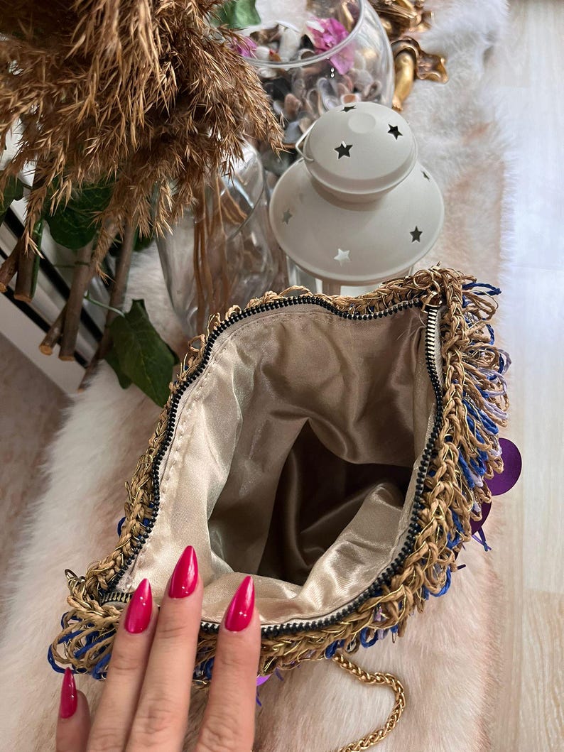 May include: Open view of a gold-colored chain bag with blue and gold fringe. The bag has a gold-colored chain strap and a beige satin interior. The zipper is black. The bag is on a white fur-like surface.