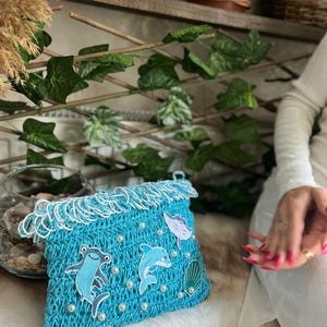May include: A turquoise crochet clutch with white fringe and pearl accents. The clutch features appliqu&eacute;s of a hammerhead shark, a dolphin, a stingray, and a seashell. The bag is designed with a marine theme.