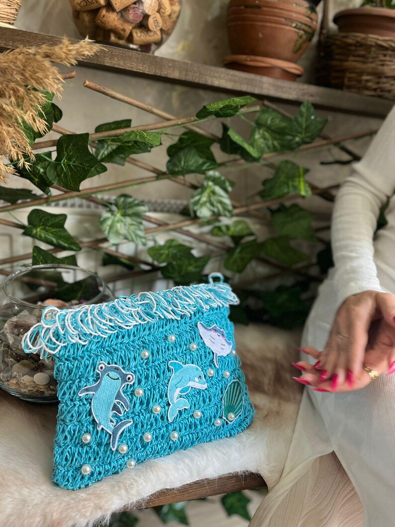 May include: A turquoise crochet clutch with white fringe and pearl accents. The clutch features appliqu&eacute;s of a hammerhead shark, a dolphin, a stingray, and a seashell. The bag is designed with a marine theme.