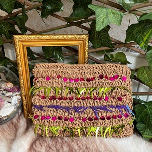 May include: A rectangular, woven clutch in a natural tan color, adorned with rows of pink sequins, green fringe, and purple stitching. The clutch is decorated with a floral design. A gold picture frame and greenery are in the background.