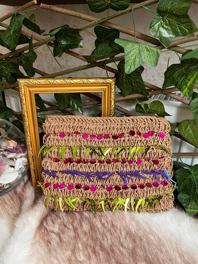 May include: A rectangular, woven clutch in a natural tan color, adorned with rows of pink sequins, green fringe, and purple stitching. The clutch is decorated with a floral design. A gold picture frame and greenery are in the background.