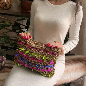 May include: A woven brown clutch with neon green, pink, and blue accents. The rectangular bag is held by a person wearing a white long-sleeved dress. The clutch features a textured design with colorful embellishments.